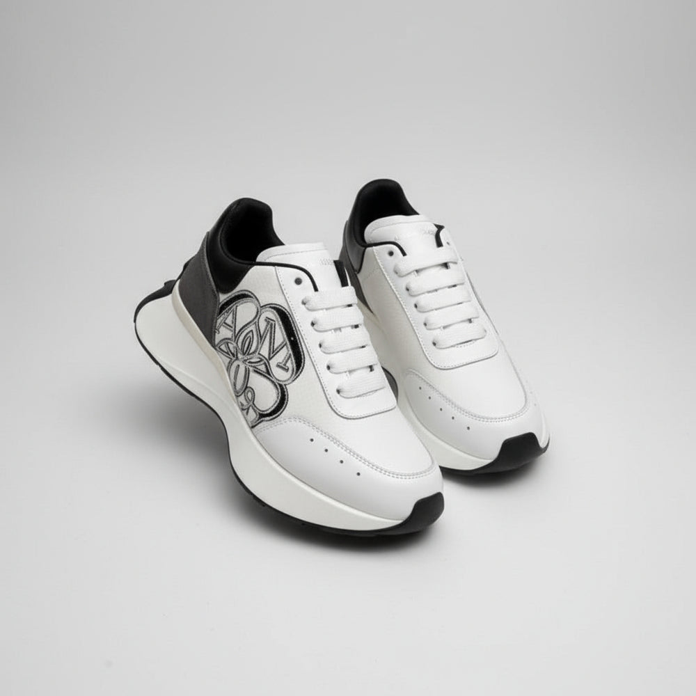 White sneakers with black accents held by a gloved hand, with Alexander McQueen boxes in the background.