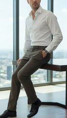 Man in a white shirt and brown pants sitting by a window with a cityscape view.