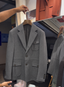 A gray double-breasted blazer with flap pockets hanging on a wooden hanger in a retail store setting, with a person's hand holding the hanger and another person partially visible in the background.