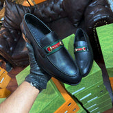 Black leather loafers with a red and green buckle held by a gloved hand on a green surface.