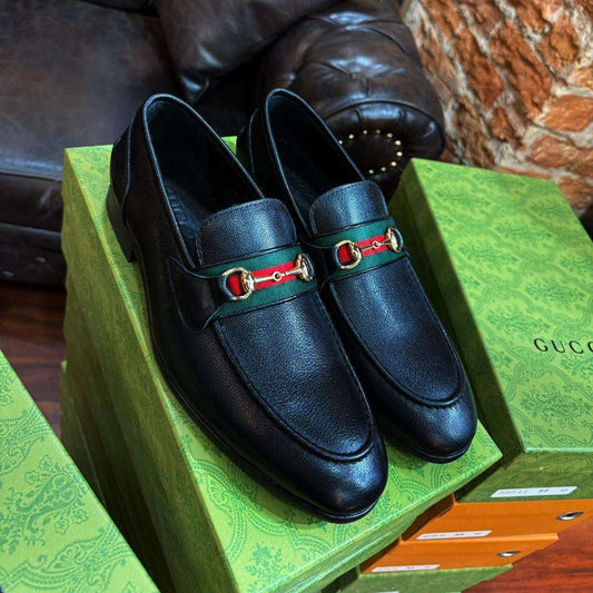 Black loafers with red and gold accents on a green Gucci box against a brick wall.