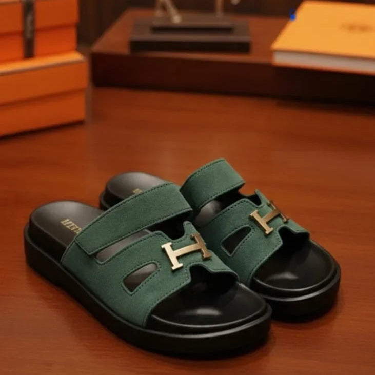 Green sandals with gold H-buckles on a wooden surface, with Hermes packaging in the background.