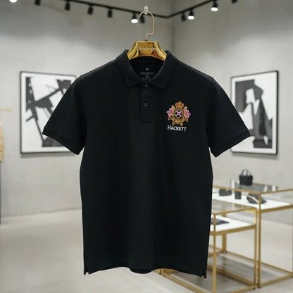 Black polo shirt with a logo on a hanger against a gray wall with framed pictures.