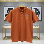 Orange polo shirt with a brand logo on a hanger against a gray wall with framed pictures.