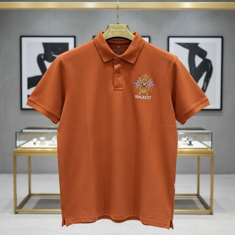 Orange polo shirt with a brand logo on a hanger against a gray wall with framed pictures.