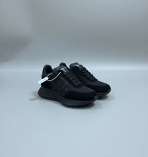 An all-black Alexander McQueen Sprint Runner sneaker being held to show its suede and leather details, embossed side logo, and thick chunky sole.