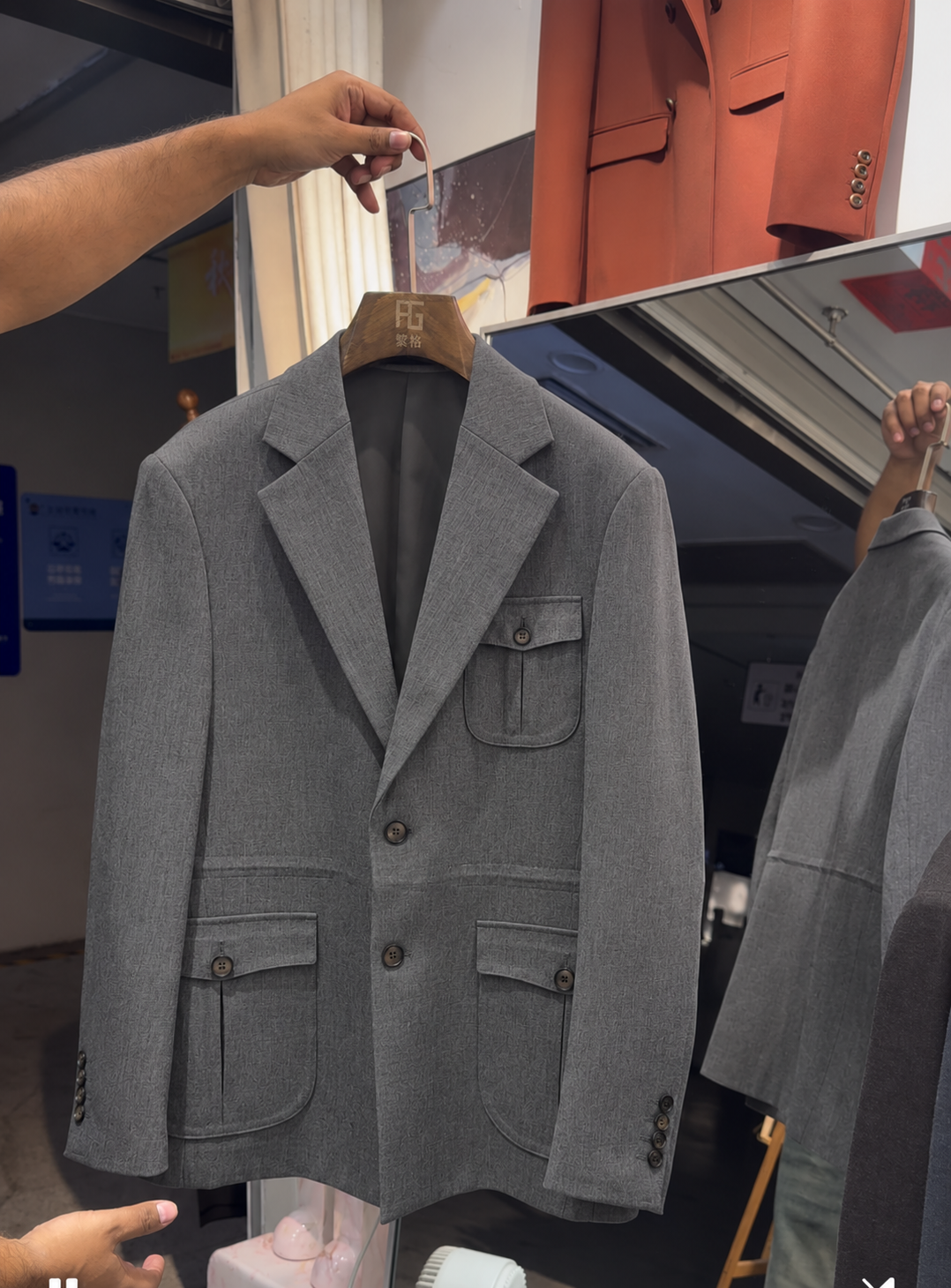 A gray double-breasted blazer with flap pockets hanging on a wooden hanger in a retail store setting, with a person's hand holding the hanger and another person partially visible in the background. Main image