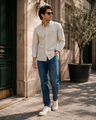 A man wearing a cream and navy vertically striped long-sleeve button-down shirt, blue jeans, and white sneakers stands on a sunlit stone sidewalk next to a building.