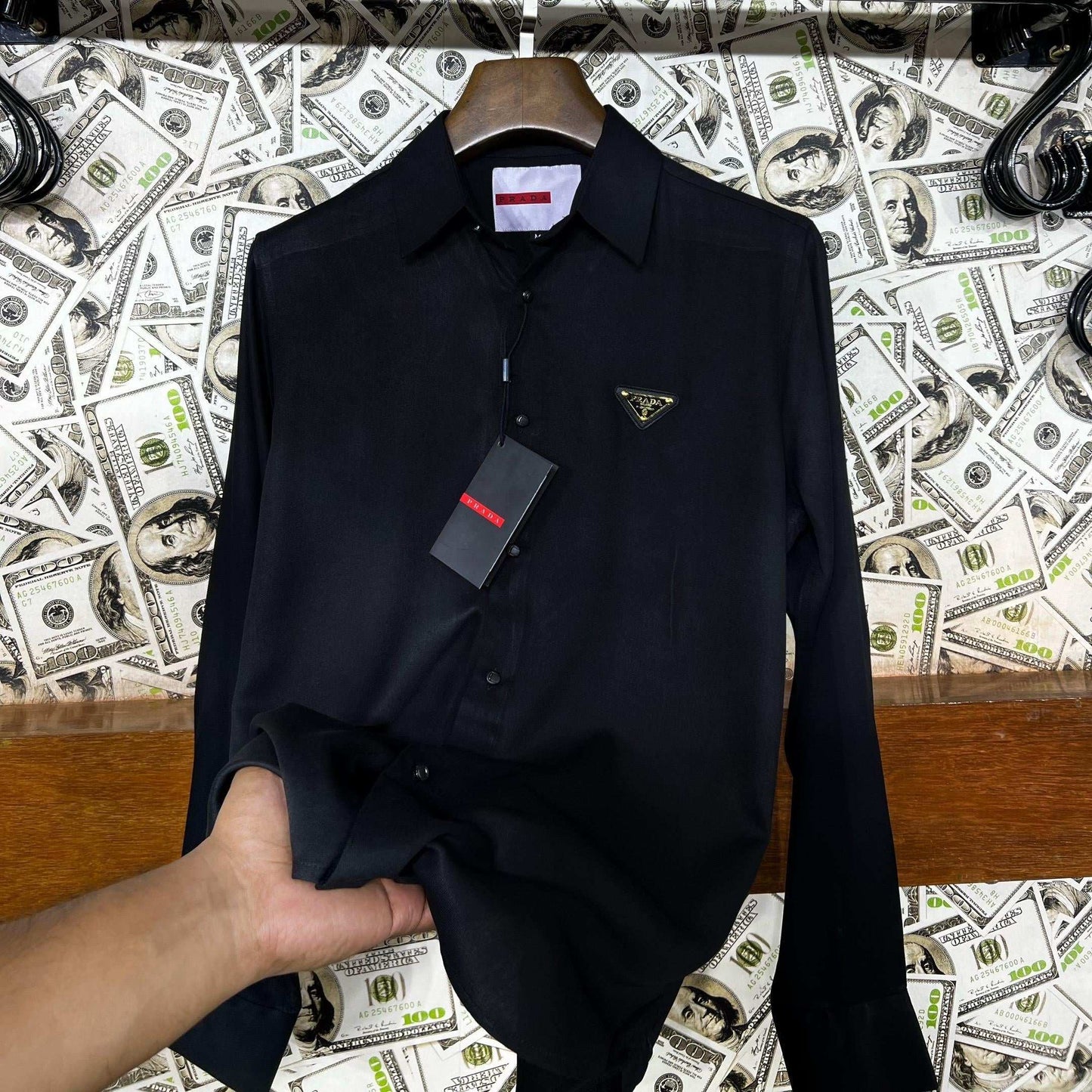 Black shirt on a hanger with a Prada tag against a background of dollar bills and 'UNIVERSES' text.