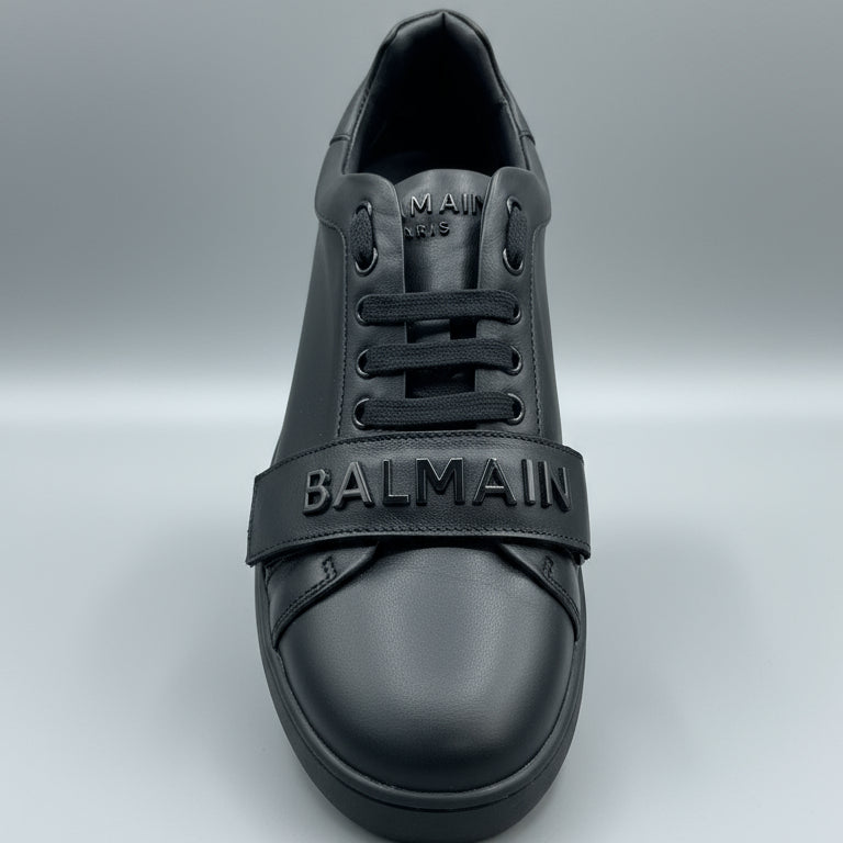 Black Balmain sneaker held in a hand with a wooden surface and dark leather couch in the background. Main image