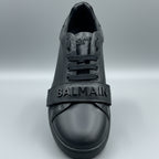 Black Balmain sneaker held in a hand with a wooden surface and dark leather couch in the background.