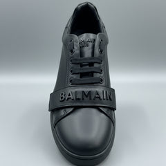 Black Balmain sneaker held in a hand with a wooden surface and dark leather couch in the background.