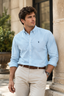 A man wearing a light blue long-sleeve button-down shirt by Polo Ralph Lauren, paired with beige trousers and a brown belt, standing outdoors next to a stone column.