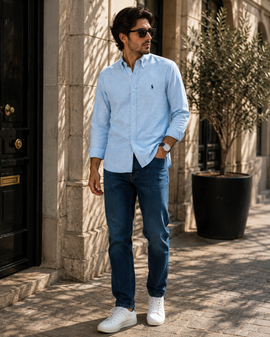 A man wearing a light blue long-sleeve button-down shirt, dark blue jeans, and white sneakers stands on a sunlit stone sidewalk next to a black door and a potted tree.