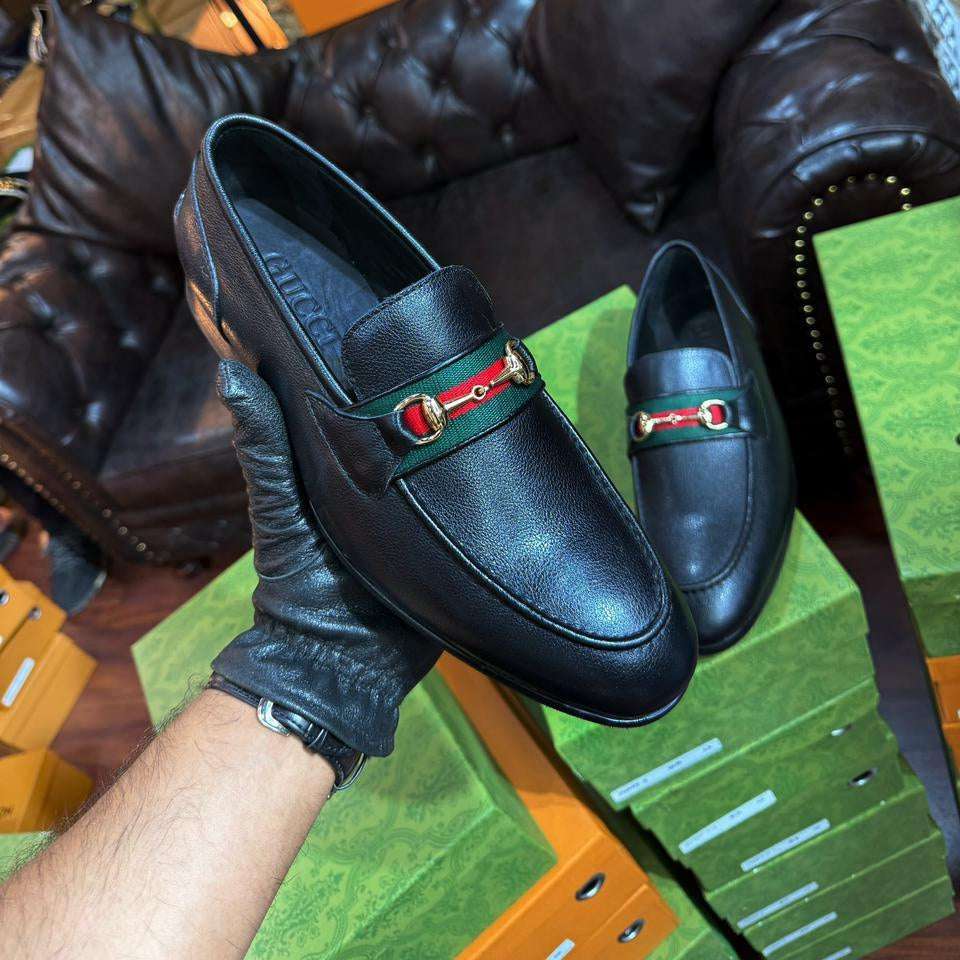 Black leather loafers with a red and green buckle held by a gloved hand on a green surface.