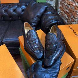 Pair of black crocodile-patterned loafers on an orange Louis Vuitton box with a black leather couch and brick wall in the background.
