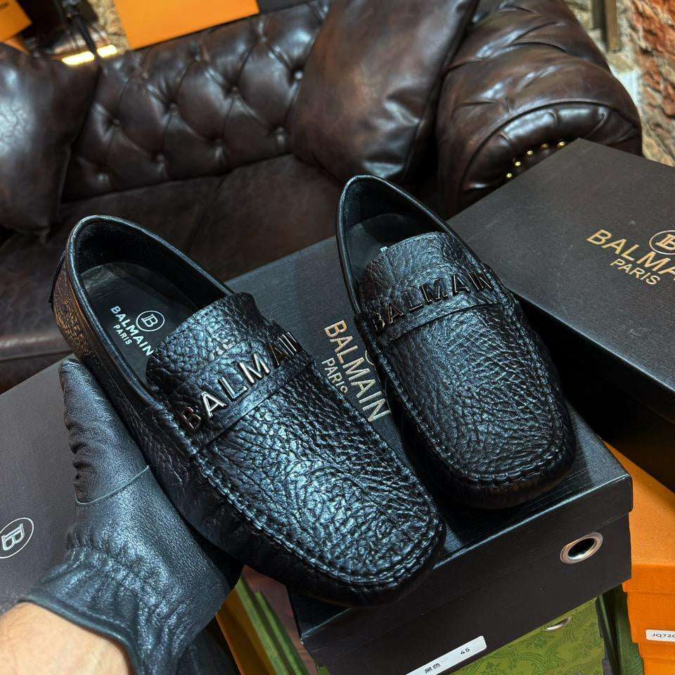 Black leather loafers with Balmain branding on a matching box against a textured wall background.