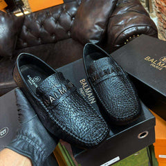 Black leather loafers with Balmain branding on a matching box against a textured wall background.