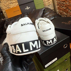 White sneakers with 'Balmain' branding held by a gloved hand against a brick wall background.