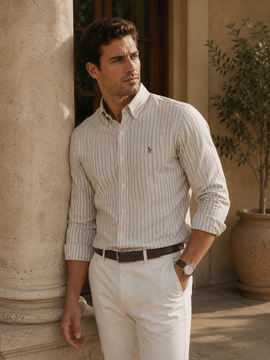 A man wearing a cream and tan vertically striped long-sleeve button-down shirt with white trousers, leaning against a stone column in an outdoor patio setting.