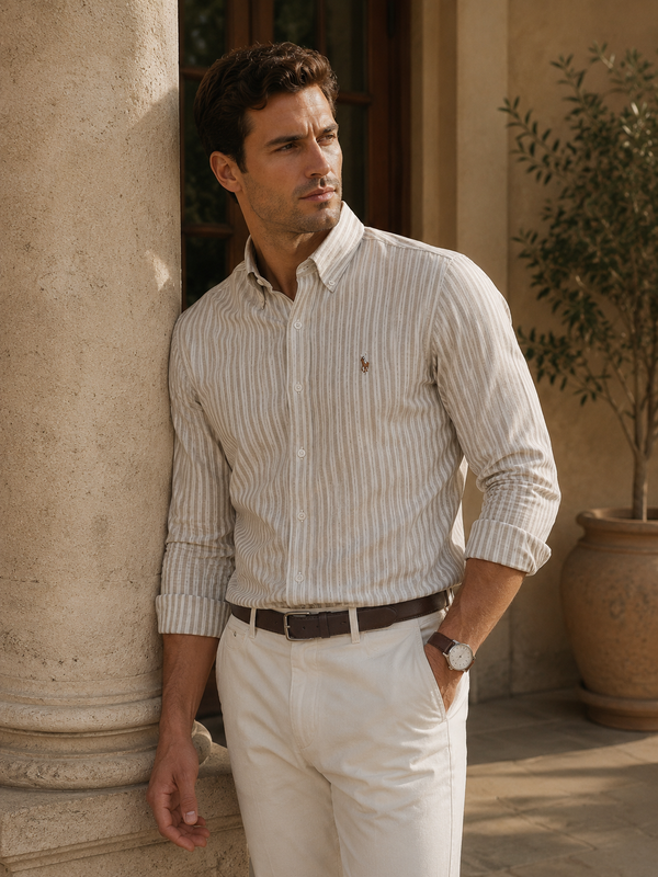 A man wearing a cream and tan vertically striped long-sleeve button-down shirt with white trousers, leaning against a stone column in an outdoor patio setting.