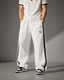 A person wearing white Puma track pants with black side stripes, a white Puma polo shirt, and black-and-white sneakers, standing against a gray studio background.