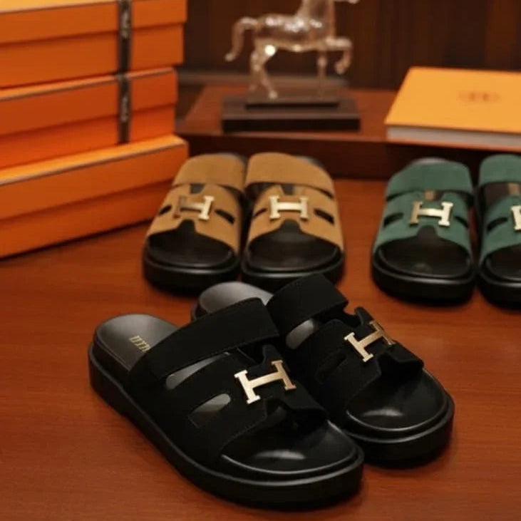 Black sandals with gold buckles on a wooden surface, with orange boxes and a horse statue in the background.