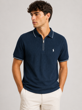 A man wearing a navy blue ribbed short-sleeve polo shirt with a half-zip collar and white piping, paired with off-white trousers, against a light gray studio background.