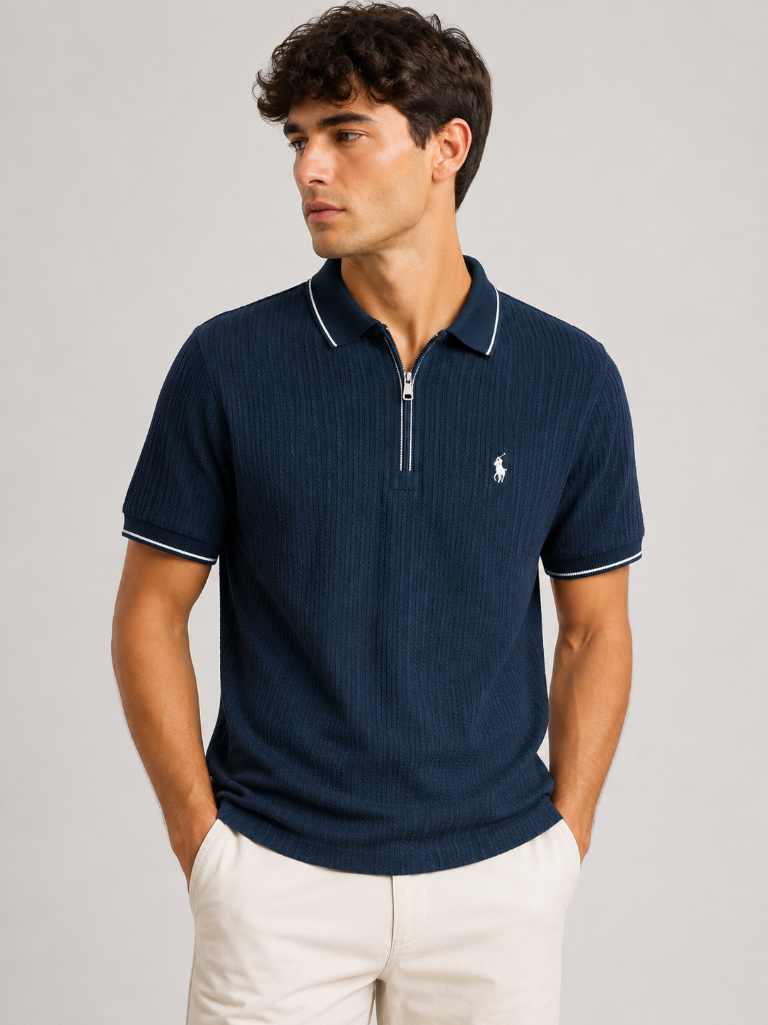 A man wearing a navy blue ribbed short-sleeve polo shirt with a half-zip collar and white piping, paired with off-white trousers, against a light gray studio background.