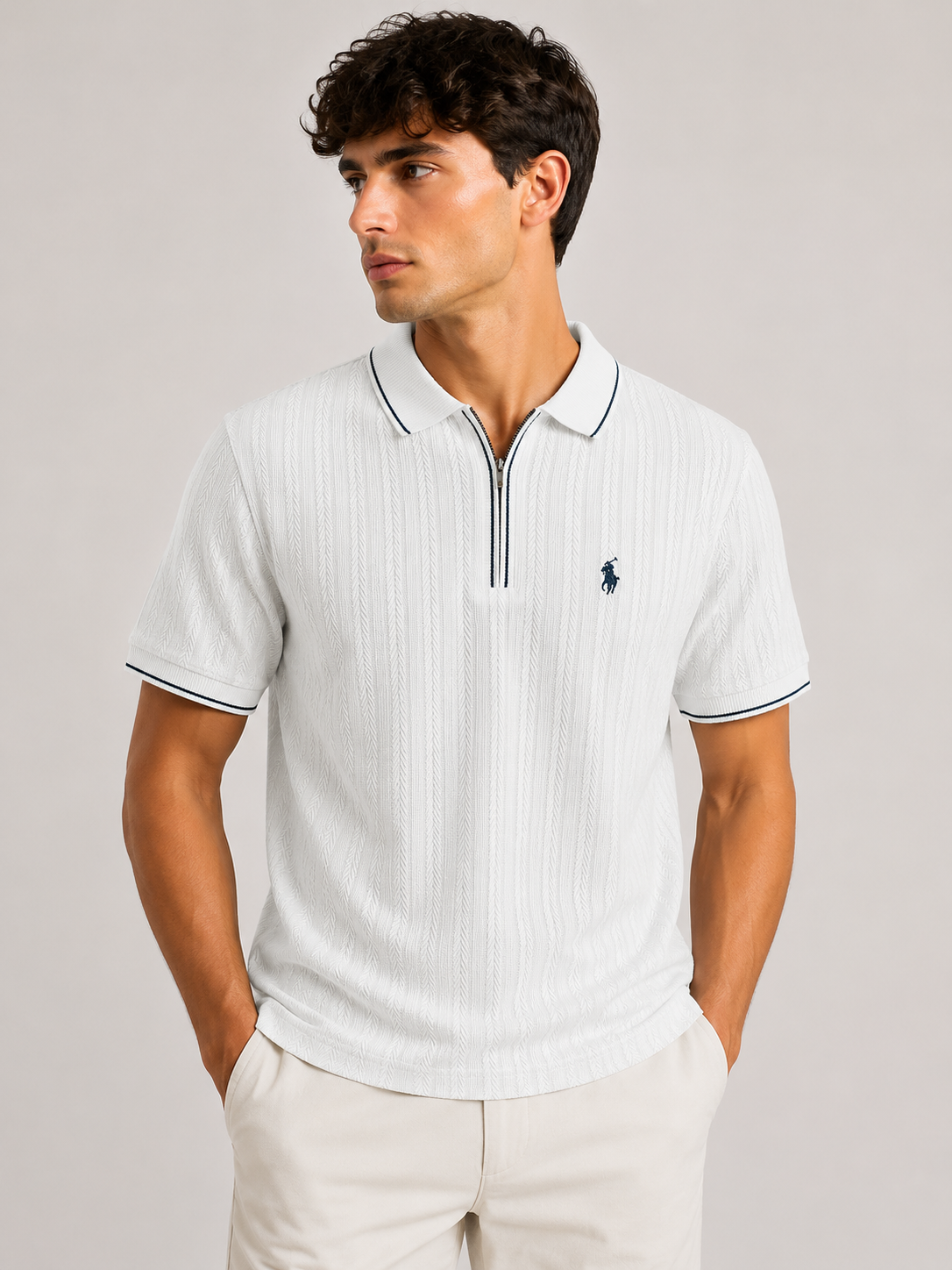 A man wearing a white short-sleeve polo shirt with thin navy blue piping and a small navy embroidered polo player logo on the chest, paired with light beige trousers, against a solid light gray background.
