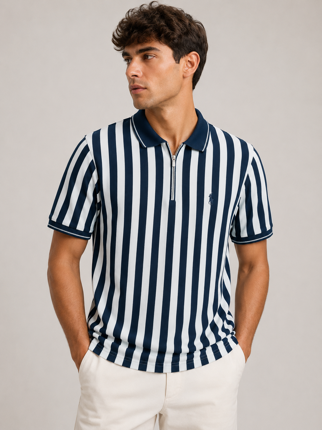 A man wearing a navy and white vertically striped short-sleeve polo shirt with a half-zip collar, paired with off-white trousers, against a plain light gray studio background.