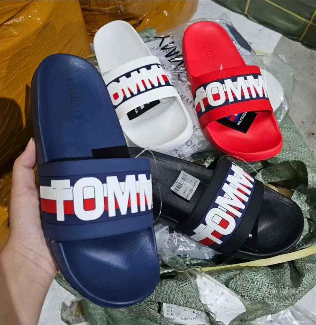Tommy Hilfiger slides in blue, white, red, and black held by a hand on a textured surface.
